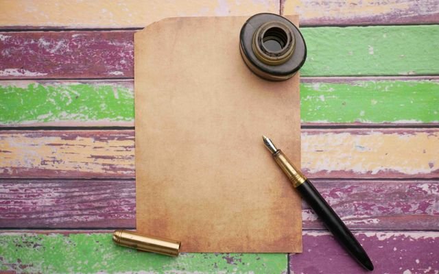 Flat lay of vintage paper with ink bottle and pen on a colorful wooden surface.