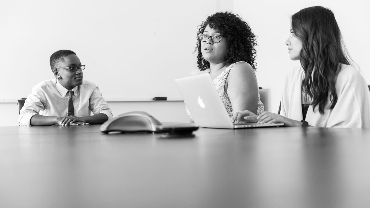 Three professionals discussing work at a conference room table with laptops in a modern office.