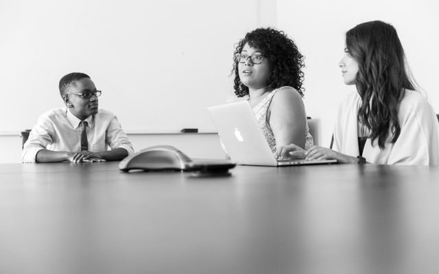 Three professionals discussing work at a conference room table with laptops in a modern office.