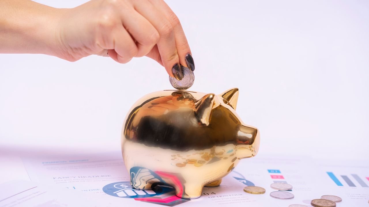 A hand places a coin into a golden piggy bank surrounded by financial documents, symbolizing savings.