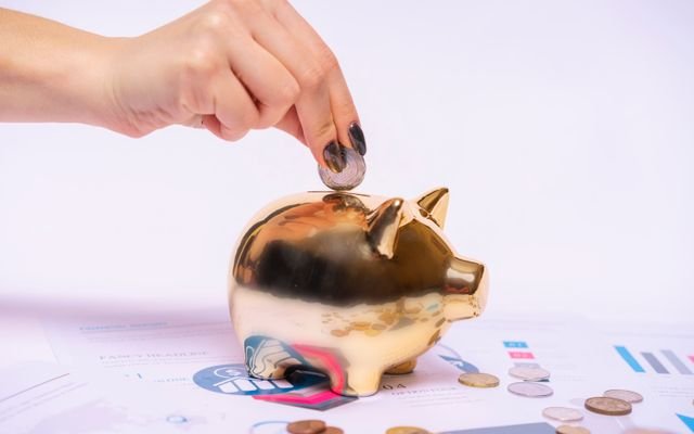 A hand places a coin into a golden piggy bank surrounded by financial documents, symbolizing savings.