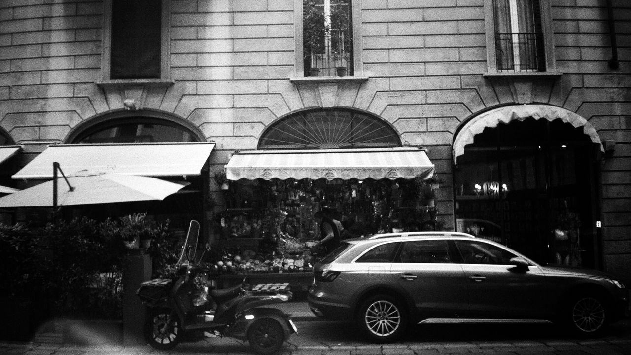 A monochrome urban street scene featuring a parked car, motor scooter, and a florist's storefront.