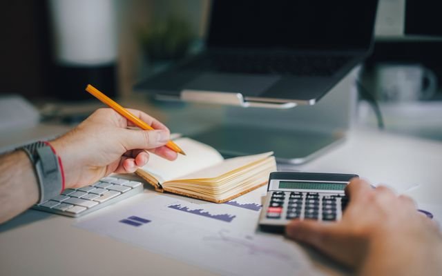 Hands using calculator and taking notes on desk with laptop, showcasing business analytics.