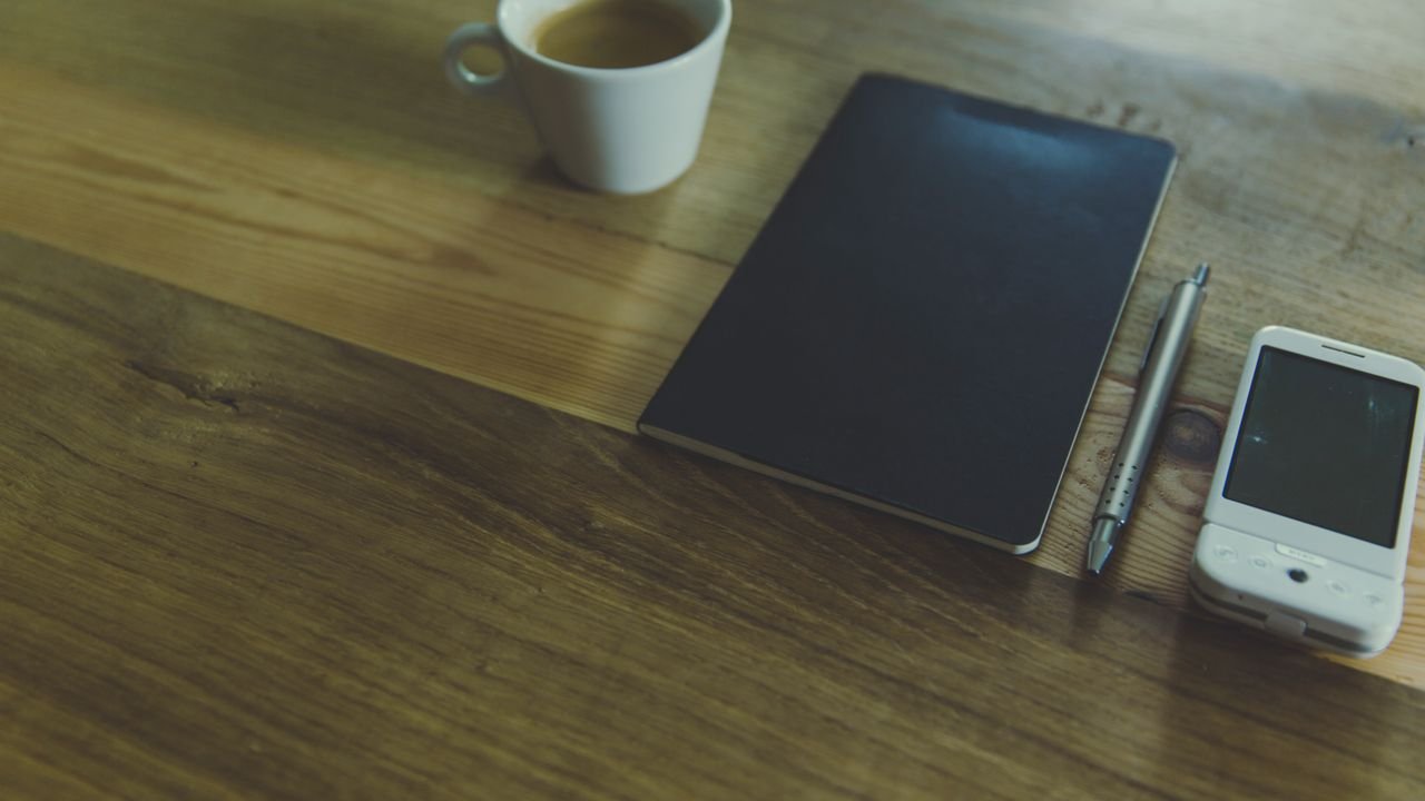 A simple workspace setup featuring a coffee cup, notebook, pen, and smartphone on a wooden desk.