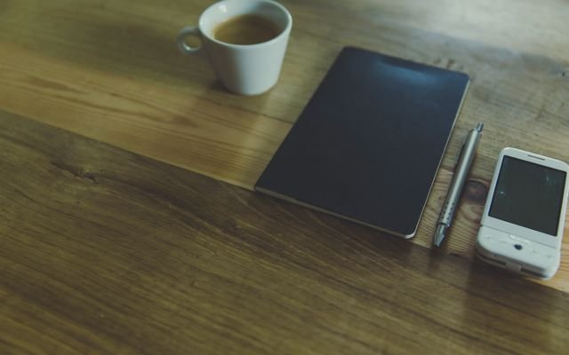 A simple workspace setup featuring a coffee cup, notebook, pen, and smartphone on a wooden desk.