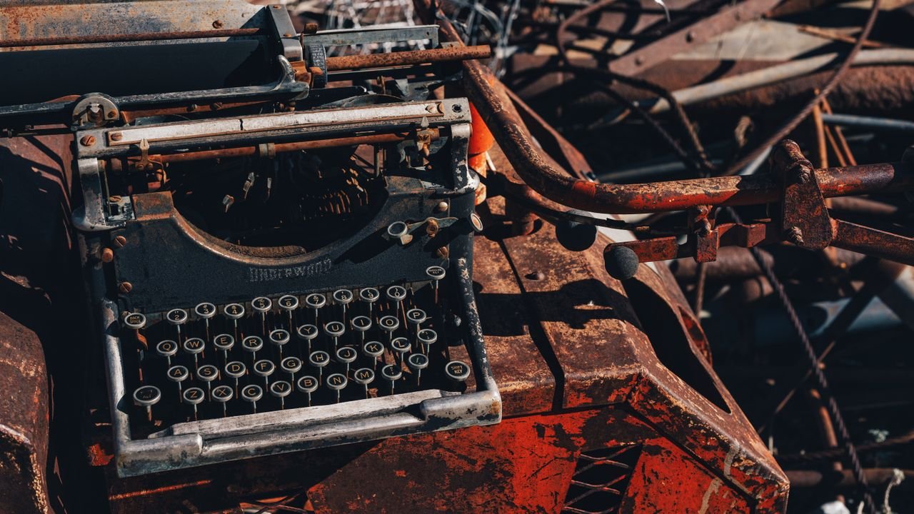 Rusty typewriter amidst metal scrap in a junkyard, bathed in harsh sunlight.