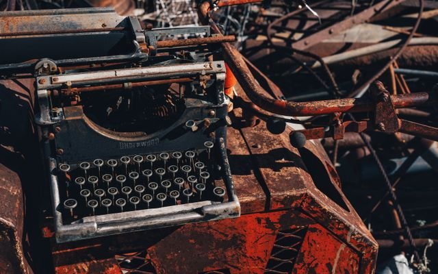 Rusty typewriter amidst metal scrap in a junkyard, bathed in harsh sunlight.