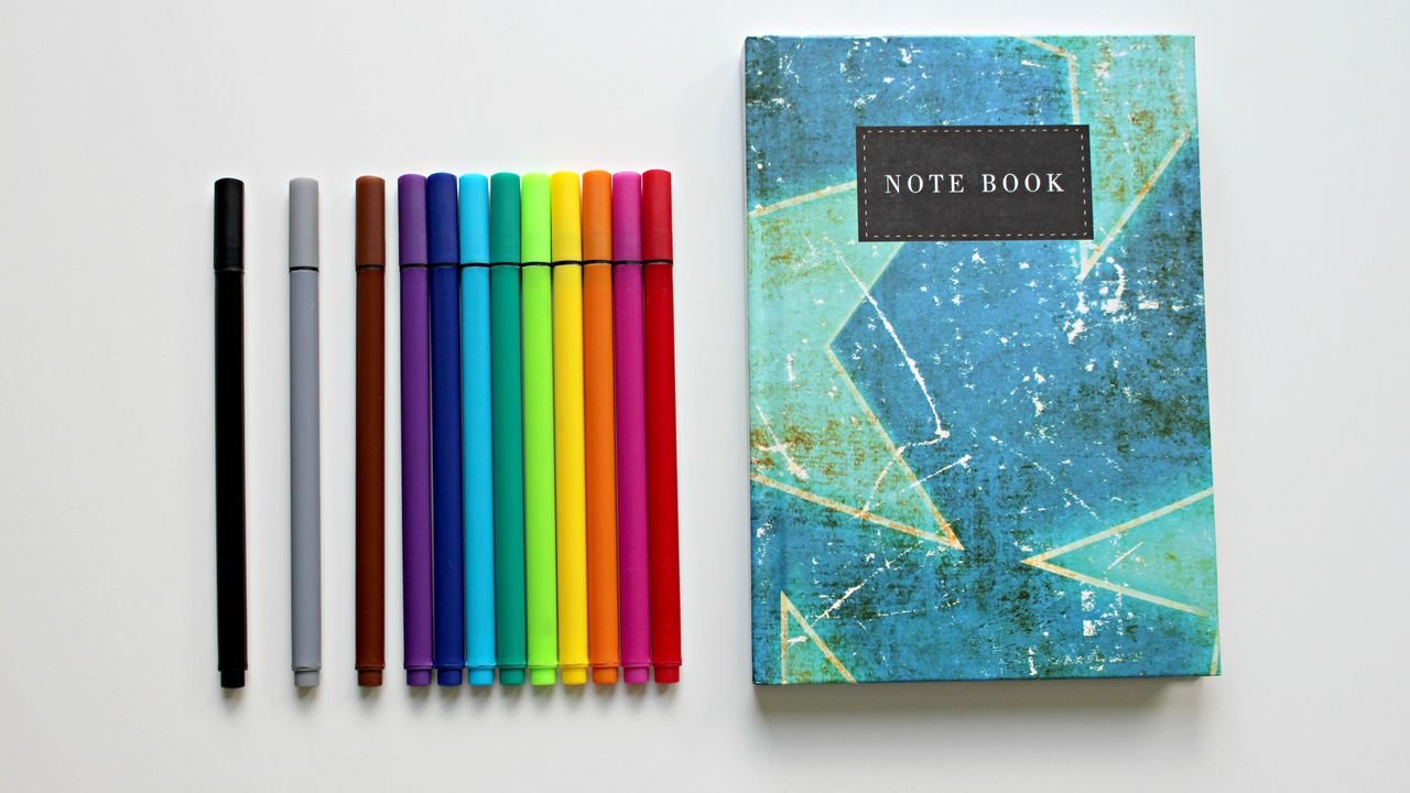 A colorful assortment of pens lined up beside a blue notebook on a white background, perfect for creative projects.