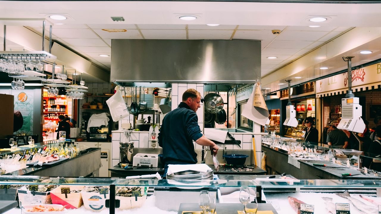 A chef prepares fresh seafood for sale at an indoor market, surrounded by ingredients and displays.