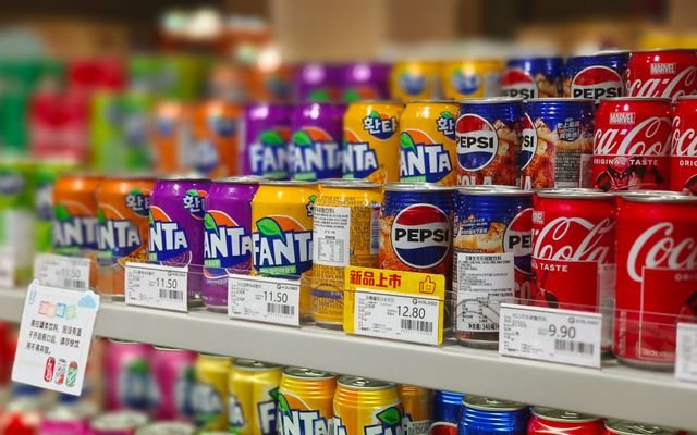 Colorful soda cans including Fanta, Pepsi, and Coca-Cola arranged on supermarket shelves, price tags visible.