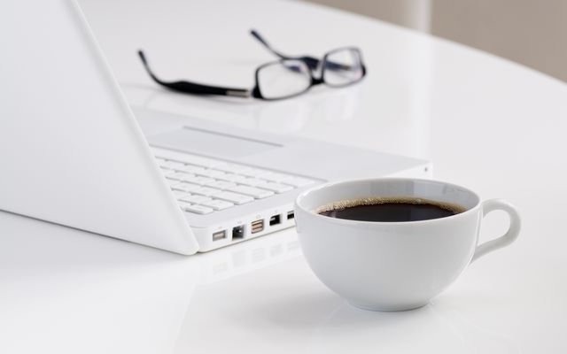 A minimalist workspace featuring a laptop, a cup of black coffee, and eyeglasses on a white desk.