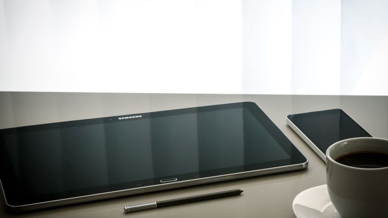 Minimalist office desk setup featuring a tablet, smartphone, pen, and coffee cup on a reflective surface.