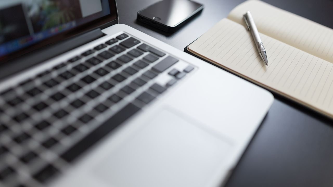 A modern workspace featuring a laptop, notebook, smartphone, and pen on a desk.