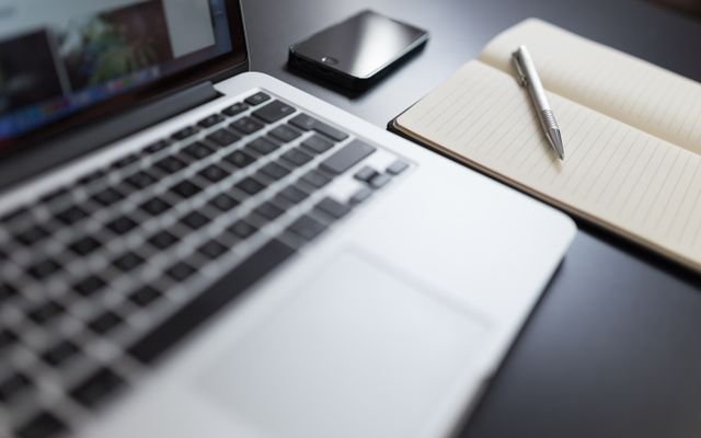 A modern workspace featuring a laptop, notebook, smartphone, and pen on a desk.