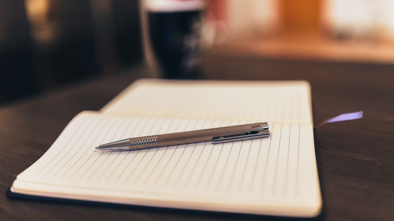 Open notebook with pen on a table, mug in background, cozy setting.