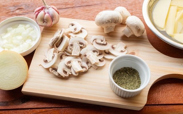 A wooden chopping board with fresh mushrooms, garlic, onion, and herbs ready for cooking.
