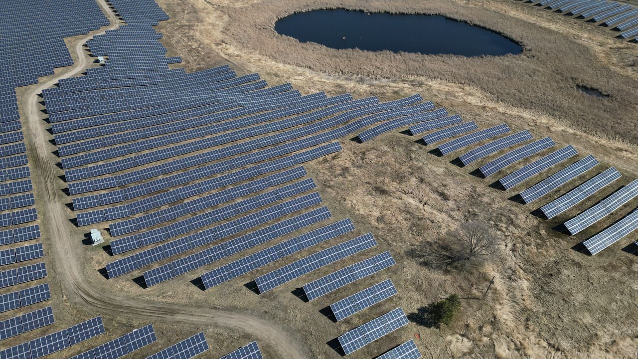 Expansive aerial view of a solar farm with rows of panels and a pond.
