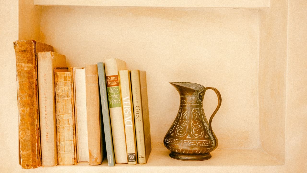 Aesthetic bookshelf with vintage books and an ornate brass jug in a warm-toned, minimalist setting.