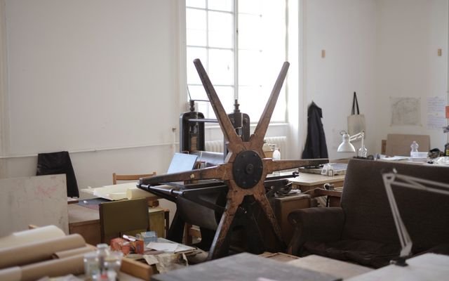 Workroom with paper rolls on table and white wall with notes and paper rolling machine with wooden mill shape wheel in afternoon