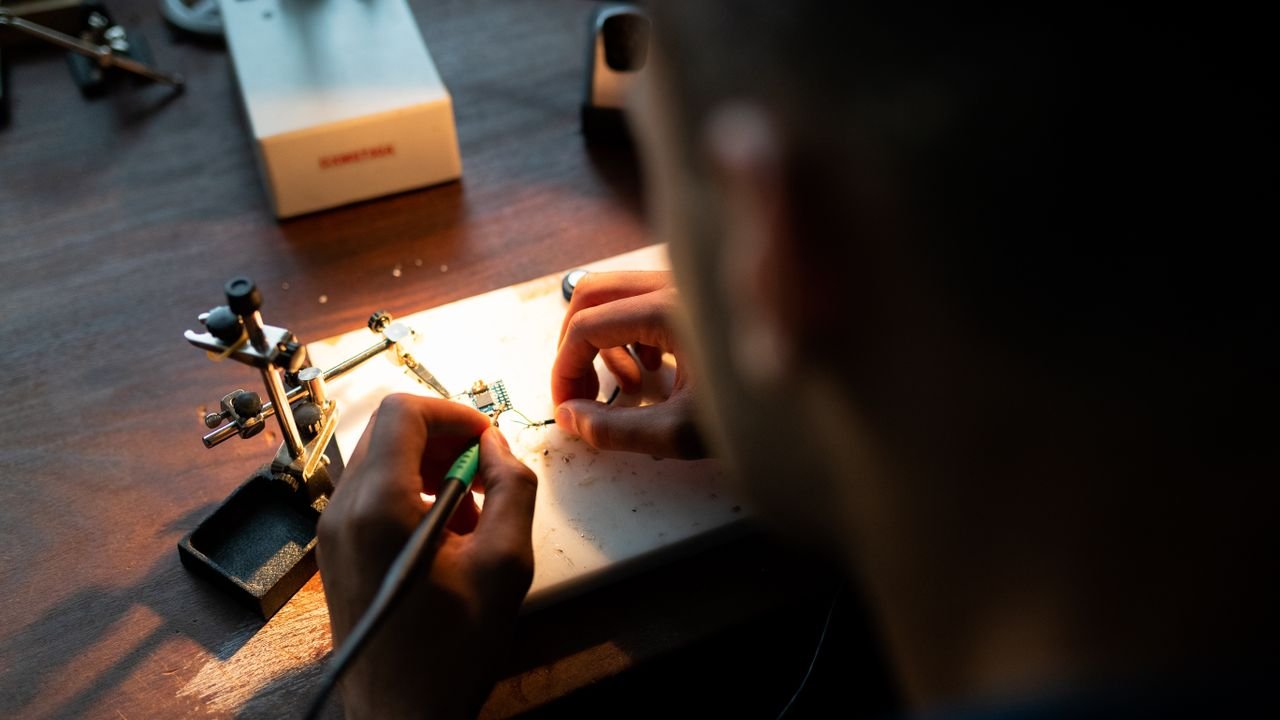 A focused engineer soldering a circuit board under bright light in a workshop setting.