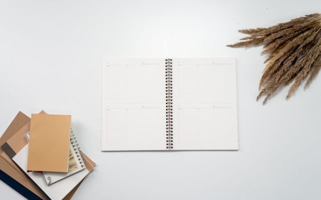 Open notebook with planner pages, surrounded by notebooks and pampas grass on a white background.