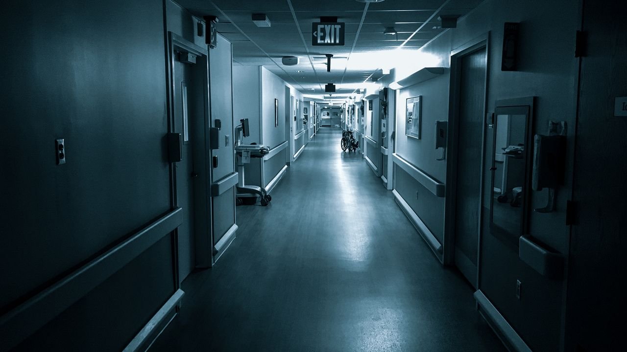 Black and white poorly lit empty wide corridor of hospital with doors on both sides and exit sign on ceiling