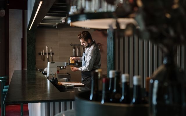 A male chef meticulously prepares a dish in a contemporary restaurant kitchen, surrounded by culinary tools.