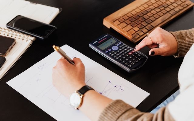 From above crop unrecognizable female accountant wearing stylish watches using calculator and writing down information on paper while working at desk with gadgets