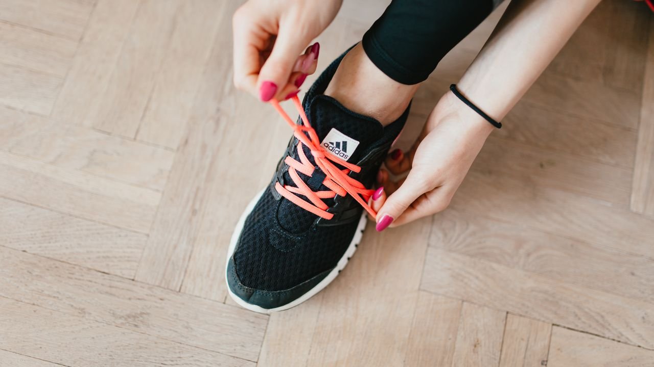 Top view of young anonymous female athlete in leggings tying shoelaces on sneakers while sitting on wooden floor in modern fitness studio and preparing for training