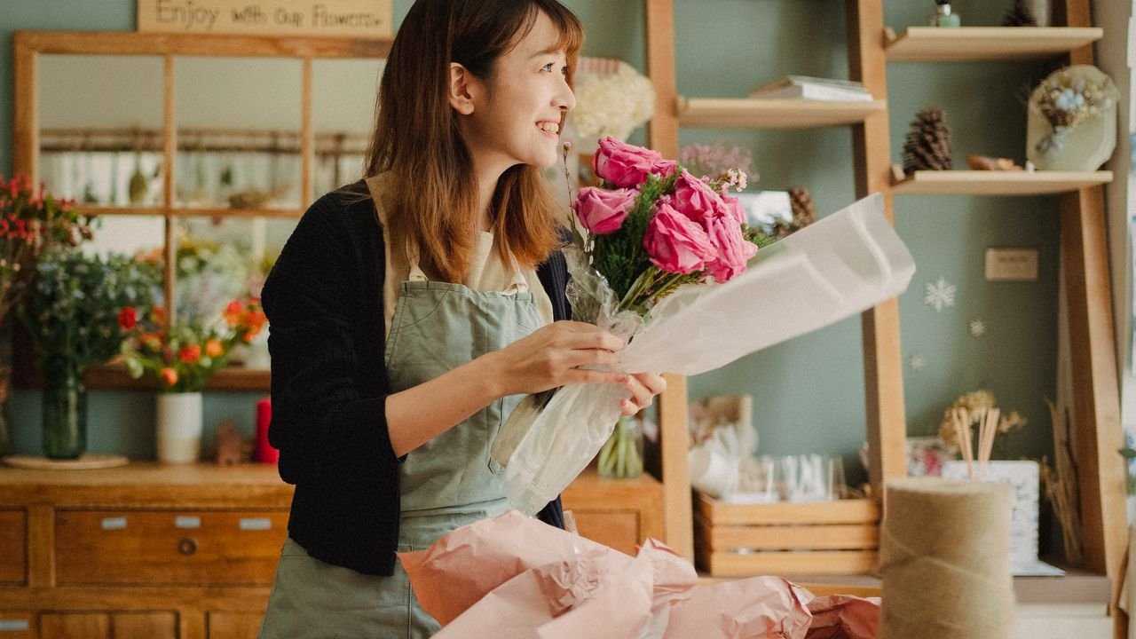 Optimistic Asian female florist wearing apron making flower bouquet with roses while standing in floral workshop