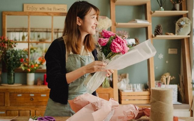 Optimistic Asian female florist wearing apron making flower bouquet with roses while standing in floral workshop