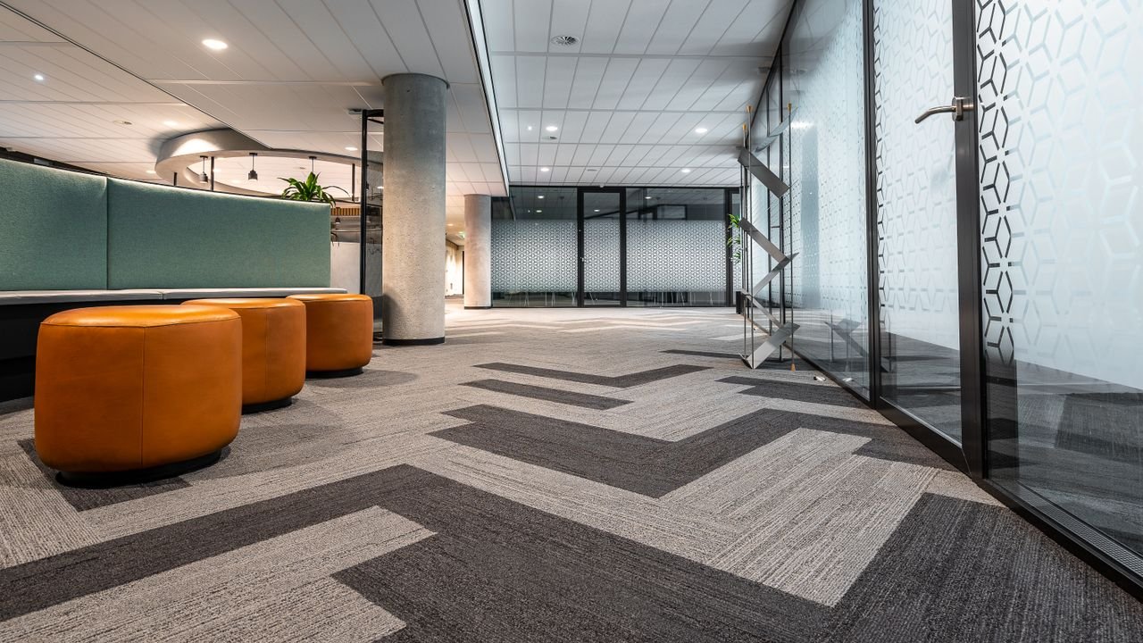 Spacious and modern office corridor featuring glass doors, geometric carpet patterns, and leather seating.