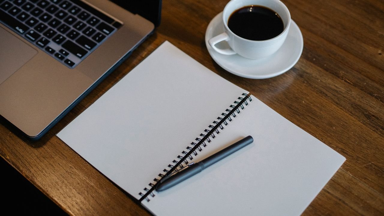 From above of workspace with cup of hot coffee and pen on empty notepad near modern laptop on wooden table