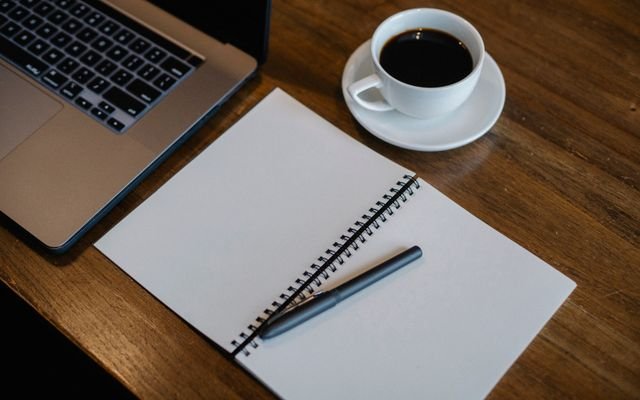 From above of workspace with cup of hot coffee and pen on empty notepad near modern laptop on wooden table