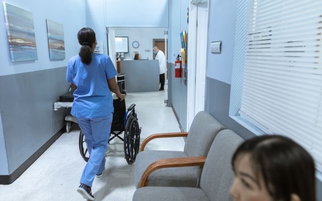 A nurse in scrubs pushes a wheelchair through a well-lit hospital corridor, attending to patient care.