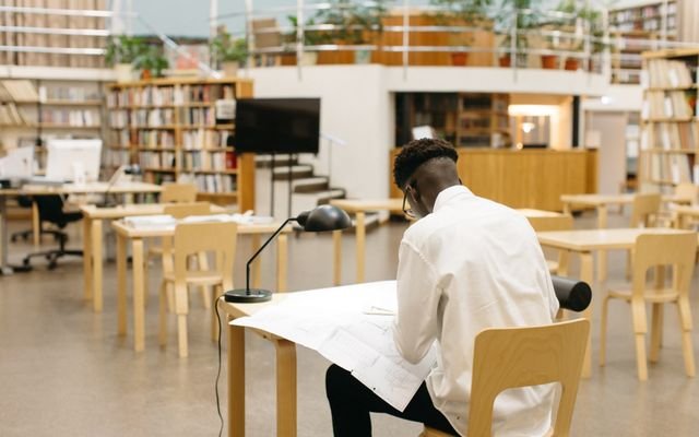 A student studying at a desk in a modern library with bookshelves and open space.