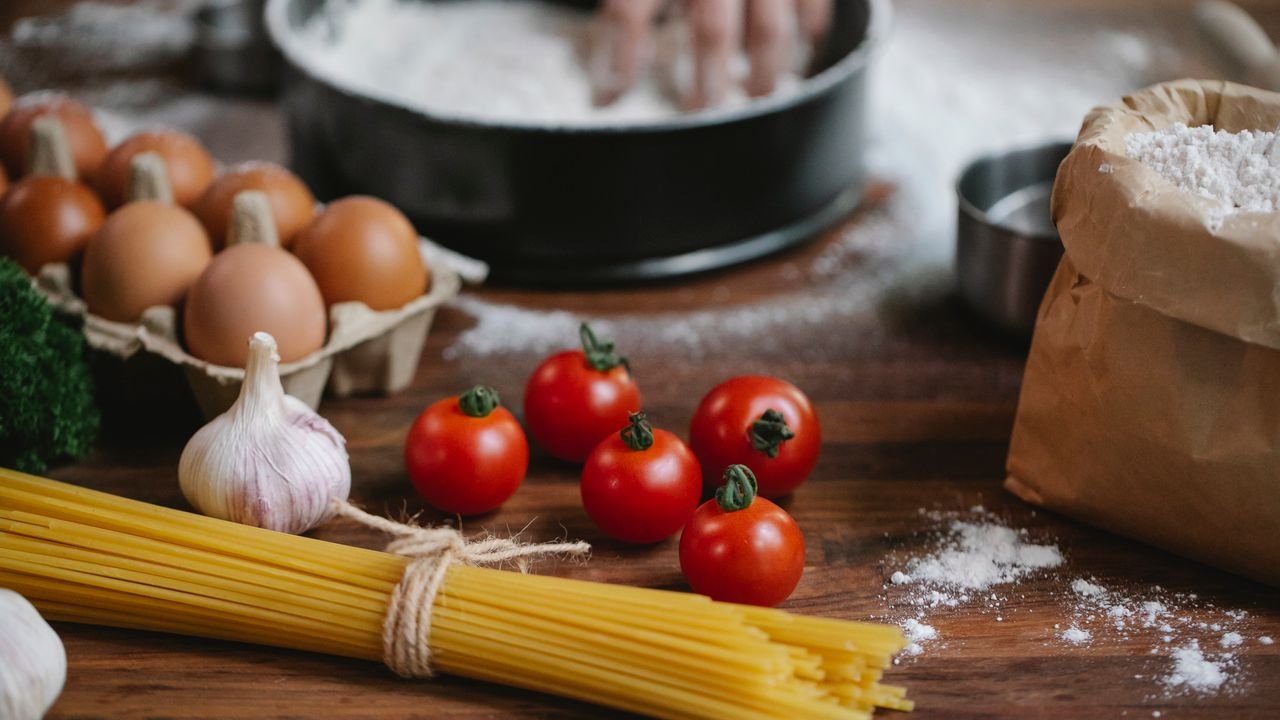 High angle of cherry tomatoes eggs garlic and spaghetti on table with crop chef kneading dough in baking dish