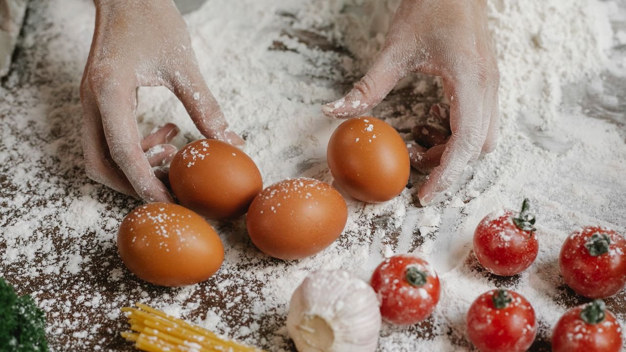 From above of crop anonymous female cook taking eggs while preparing meal with cherry tomatoes garlic spaghetti and flour