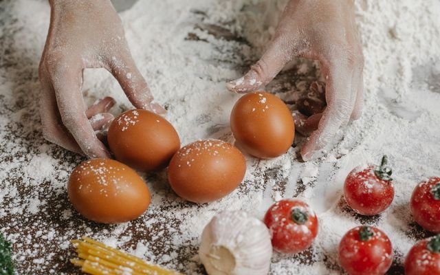 From above of crop anonymous female cook taking eggs while preparing meal with cherry tomatoes garlic spaghetti and flour