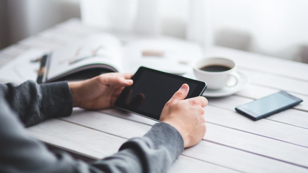 Adult holding a tablet at a minimalist workspace with coffee and a smartphone nearby.