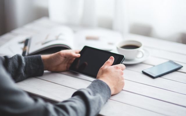 Adult holding a tablet at a minimalist workspace with coffee and a smartphone nearby.