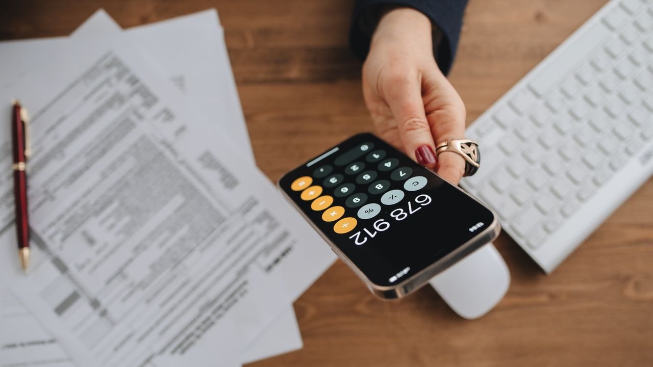 Hand holding smartphone calculator over tax documents on desk.