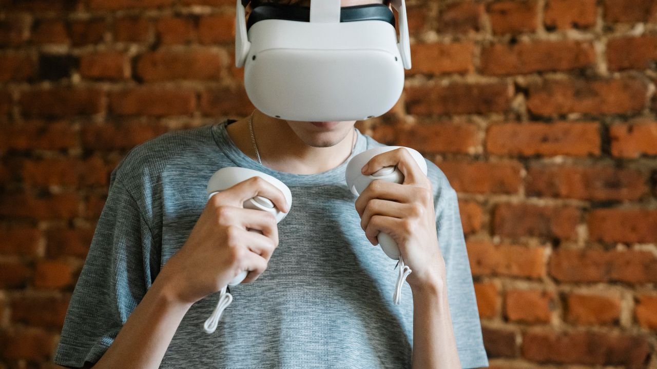 Young unrecognizable male in gray casual t shirt experiencing virtual reality with headset and controllers on background of brick wall