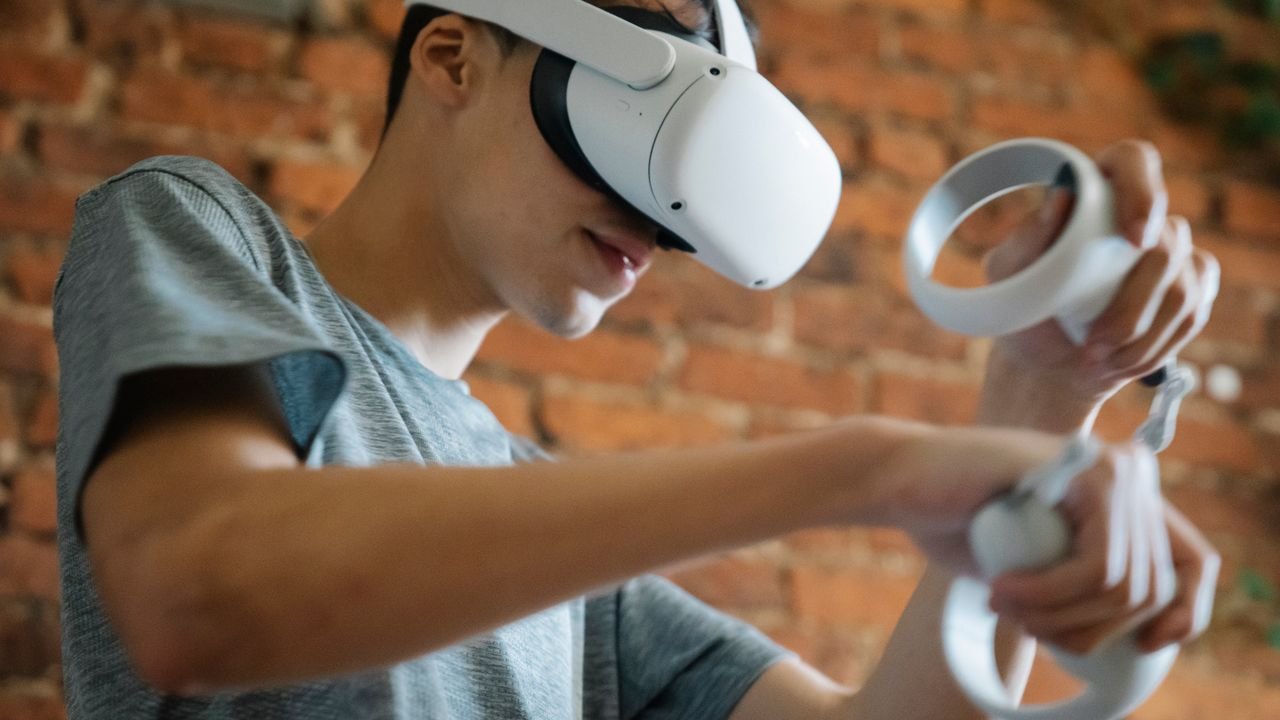 Crop young male in casual clothes using goggles of virtual reality and controllers while standing near brick wall