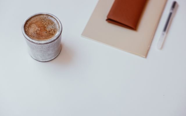 A minimalist setup with a cup of coffee, a notebook, and a pen on a white tabletop.