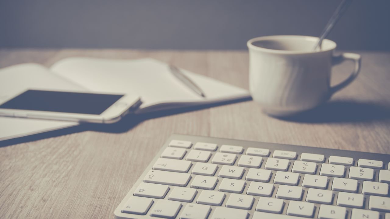 Cozy home office setup with keyboard, smartphone, notebook, and coffee cup on a wooden desk.