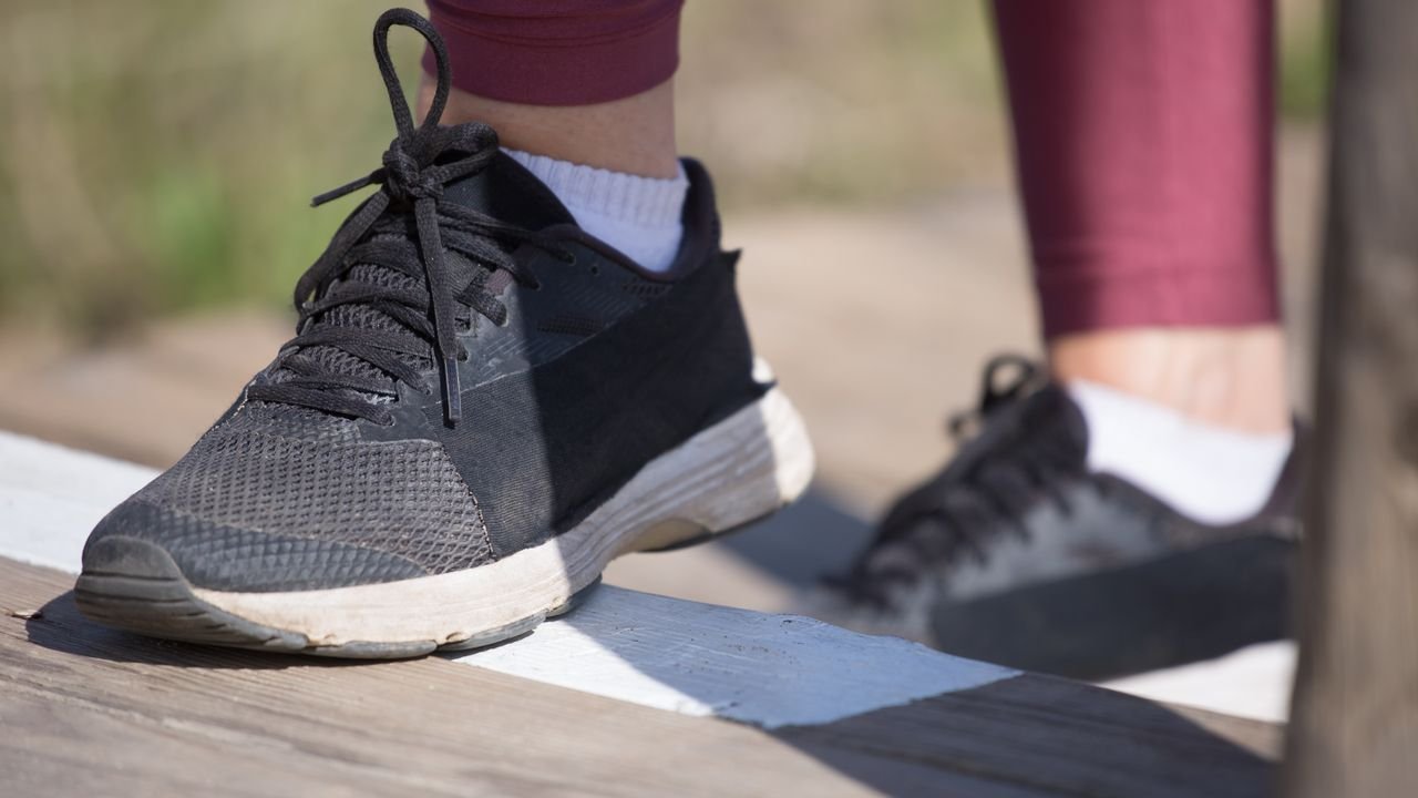 A close-up of black athletic shoes stepping onto outdoor wooden stairs in daylight.
