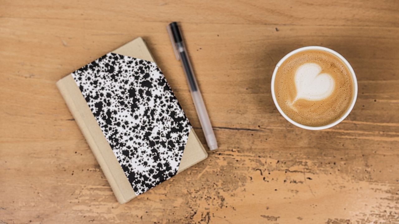 A comforting flatlay featuring a latte, marble notebook, and pen on a wooden table.