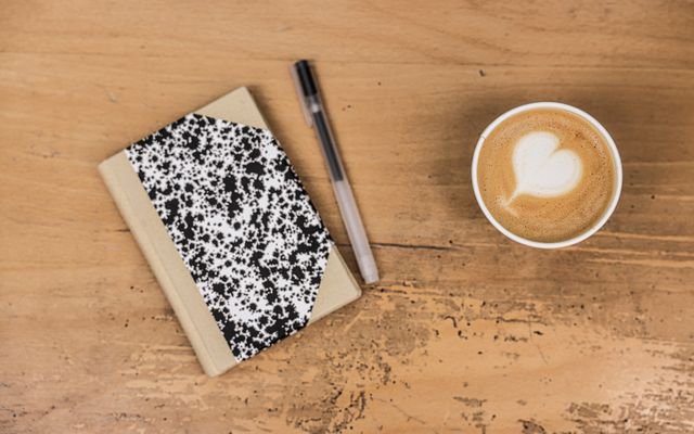 A comforting flatlay featuring a latte, marble notebook, and pen on a wooden table.
