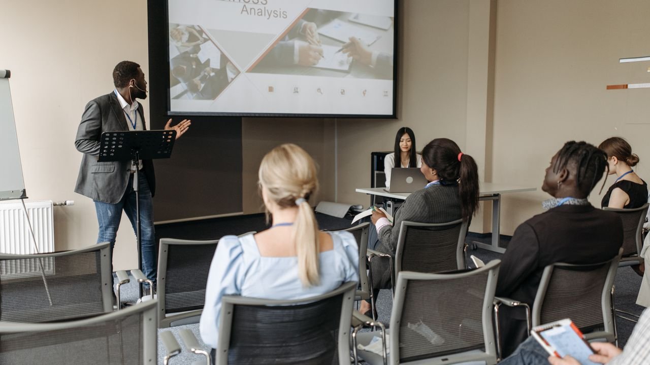 A diverse group attends a business analysis seminar with a presenter and projection screen in a modern conference room.